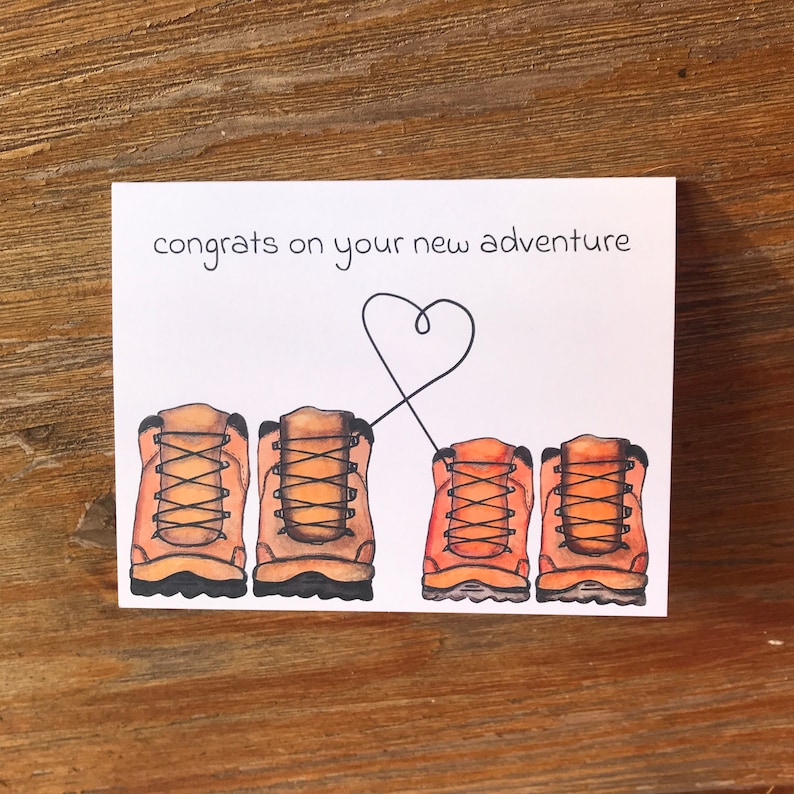 May include: A white card with the text "congrats on your new adventure" and a heart drawn with a black line. Four brown hiking boots are drawn below the text, with the two on the left connected by the heart line.