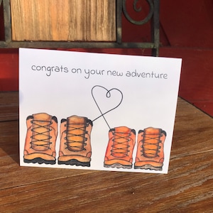 May include: A white card with the text "congrats on your new adventure" and a heart drawn with a line connecting two pairs of brown hiking boots.