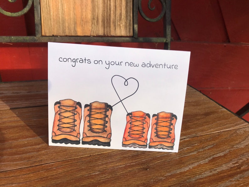 May include: A white card with the text "congrats on your new adventure" and a heart drawn with a line connecting two pairs of brown hiking boots.