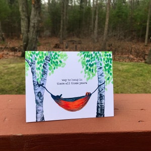 May include: A white card with a watercolor illustration of a hammock hanging between two birch trees. The hammock is red and orange and has a person lying in it. The text on the card reads "way to hang in there all those years".