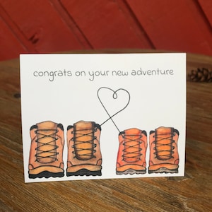 May include: A white card with the text "congrats on your new adventure" and a heart drawn with a black line. There are four brown hiking boots with orange laces drawn on the card.