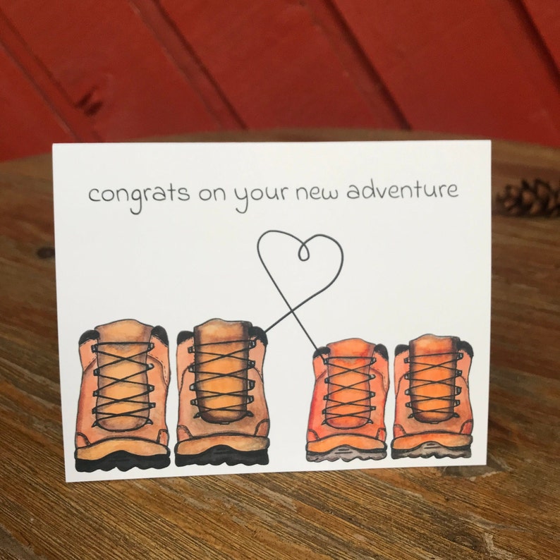 May include: A white card with the text "congrats on your new adventure" and a heart drawn with a black line. There are four brown hiking boots with orange laces drawn on the card.