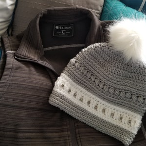 May include: A gray crocheted beanie with a white pom pom and a white band with silver embellishments. The beanie is lying on a gray and brown striped fabric.