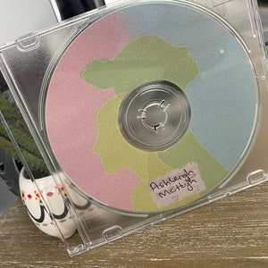 May include: A clear plastic CD case with a pink, green and blue CD inside. The CD has a silver centre and a handwritten label that says "Ashleigh McHugh".