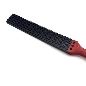 May include: A long, rectangular tool featuring a black textured surface with a repeating tyre tread pattern. The handle section is red and includes three small circular indentations arranged in a vertical line near the connection point.
