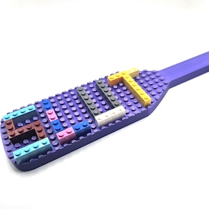 May include: A purple paddle-shaped building block base featuring a grid of studs. Attached to the surface are various small plastic bricks in light blue, brown, pink, dark blue, grey, white, and yellow, arranged to form the letters E, S, and T.