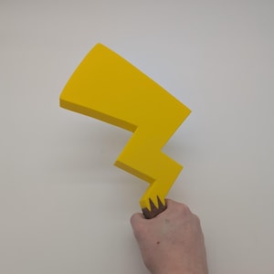 Peut inclure: Un accessoire en forme d'éclair jaune avec une poignée brune. L'accessoire est tenu dans la main d'une personne.