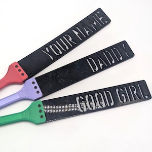 May include: Three flat, rectangular paddles with black blades and handles in red, lavender, and green. Each blade features cut-out text: 'YOUR NAME', 'DADDY', and 'GOOD GIRL'. The handles are attached to the blades with three small black screws.