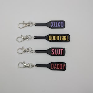 Puede incluir: Cuatro llaveros negros en forma de pala con texto bordado de diferentes colores. Los llaveros dicen "XOXO", "GOOD GIRL", "SLUT" y "DADDY".