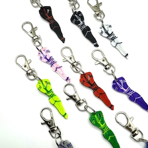 May include: A collection of ten acrylic keychains, each featuring a stylised human figure bound in rope patterns. The keychains come in various solid colours including black, white, pink, lime green, red, purple, and dark green, with silver-tone clasps.