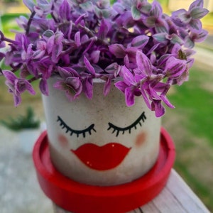 May include: A small, white ceramic planter with a painted face, featuring closed eyes and red lips. The planter is filled with purple and green foliage and sits on a red saucer.
