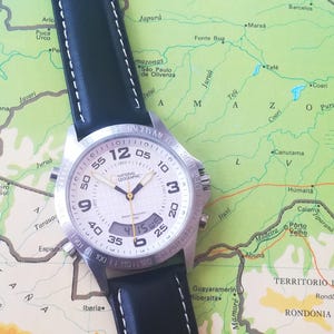 May include: A silver National Geographic watch with a black leather band and white stitching. The watch face displays the time and has a digital display. The watch is set on a colorful map.