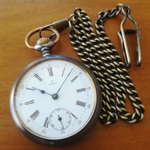 May include: A silver pocket watch with Roman numeral markings and a white face. The watch has a gold chain attached to it. The watch face says "Omega".