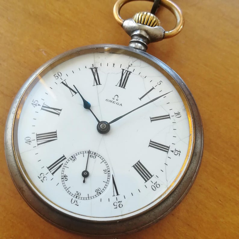 May include: A silver pocket watch with Roman numeral markings on the face. The watch has a white face with black hands and a small sub-dial. The watch is marked with the word "OMEGA".