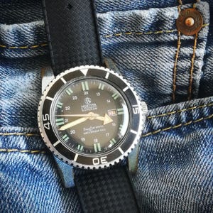 Puede incluir: Un reloj de buceo Mortima Super 20 vintage con bisel negro y correa de caucho negro. La esfera del reloj es oscura con agujas e indicadores luminosos. El reloj está sobre un fondo de vaqueros azules.
