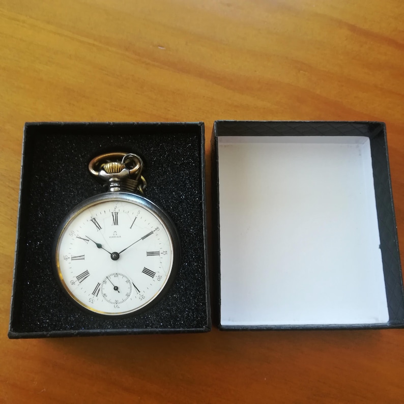 May include: A silver pocket watch with Roman numeral markings on the face, in a black velvet box. The watch has a gold-colored winding stem and a small second hand.