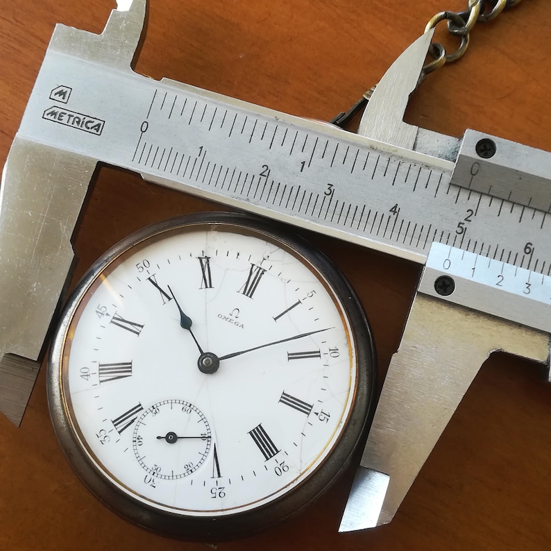 May include: A silver pocket watch with Roman numeral markings on the face. The watch is being measured with a silver caliper. The watch face reads "OMEGA" and has a second hand dial at the bottom.