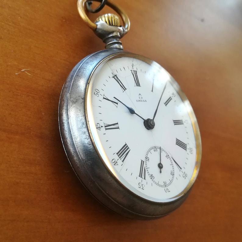 May include: A silver pocket watch with a white face and black hands. The watch has Roman numeral hour markers and a small second hand dial. The watch face says "Omega" at the top.