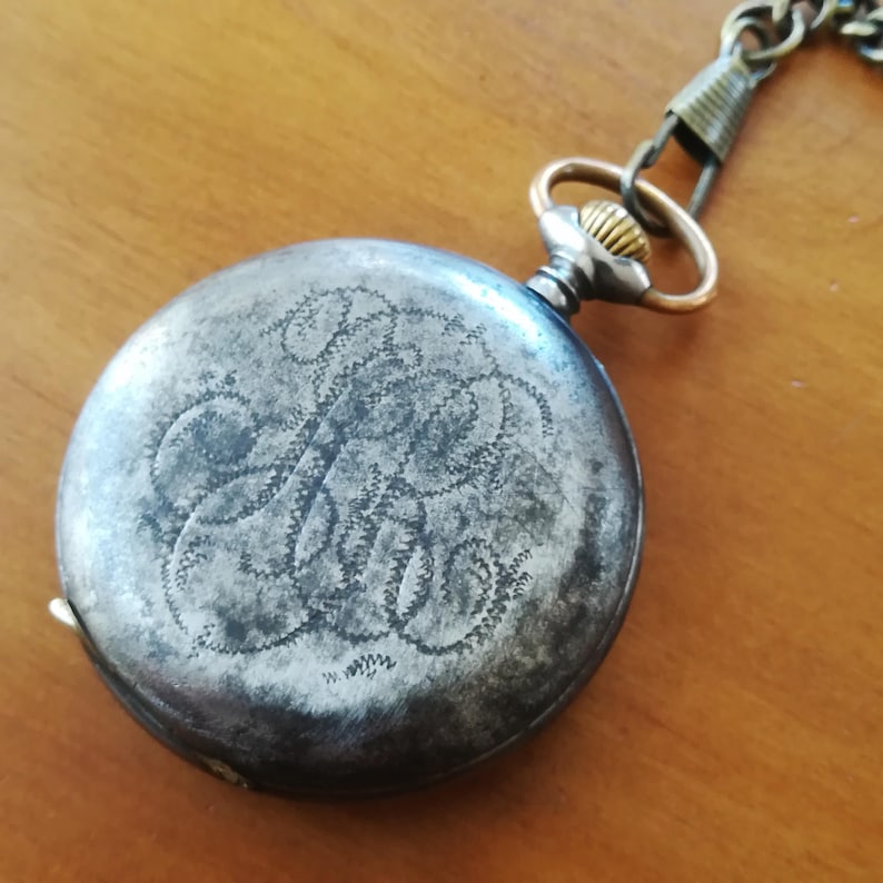 May include: A silver pocket watch with a chain attached. The watch face is engraved with a decorative pattern.