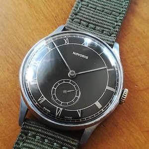 Peut inclure: Une montre-bracelet à cadran noir avec des aiguilles argentées et des chiffres romains. Le cadran de la montre porte l'inscription "NOVORIS" en blanc et les mots "SWISS MADE" en noir. La montre est munie d'un bracelet en tissu vert.