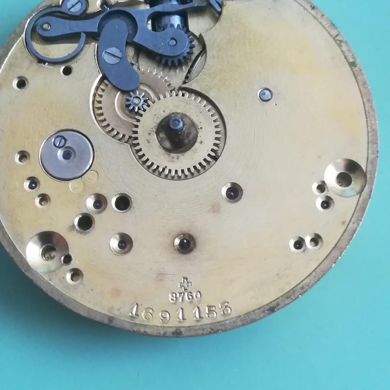 May include: Close-up of a gold-colored watch movement with intricate gears and a cross symbol. The watch movement has the number 9760 and 1691158 printed on it.
