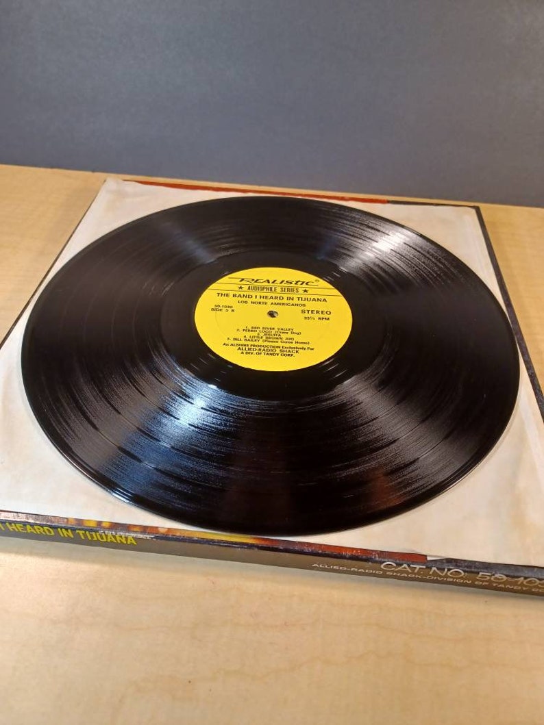 May include: A black vinyl record with a yellow label. The label says "Realistic Audiophile Series" and "The Band I Heard in Tijuana" with the band's name "Los Norte Americanos". The record is in a black sleeve with the text "The Band I Heard in Tijuana" printed on the front.