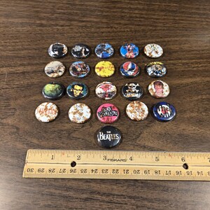 May include: A collection of vintage button pins with various designs and band logos, including The Beatles and Sex Pistols. The pins are arranged on a wooden surface with a ruler, showcasing their small size. The pins have a diameter of approximately 1 inch.