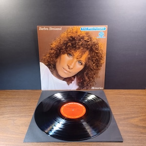 May include: A black vinyl record with a red label that says "Half Speed Mastered" and "Barbra Streisand Memories". The record is on a black mat. The album cover is in the background and features a photo of Barbra Streisand.