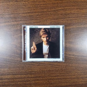 May include: A CD case featuring Daryl Hall's album "Sacred Songs." The album cover shows a portrait of Daryl Hall in a black jacket and white shirt, with the title and artist's name at the top. The CD case is in a clear plastic case.