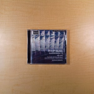 May include: A CD case with a blue and white image of a building on the front. The text on the front reads "Philip Glass Glassworlds 4 On Love The Hours Modern Love Waltz Notes on a Scandal Music in Fifths Nicolas Horvath".