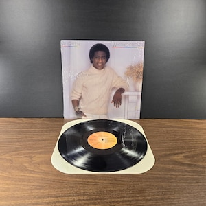 May include: A vinyl record album titled "White Christmas" by Al Green. The album cover features a photo of Al Green in a cream-colored turtleneck. The record is black with a yellow and orange label. The album is on a wooden surface.