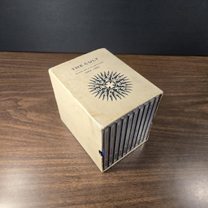 May include: A beige box set of CDs titled "THE CULT Singles Collection 1984-1990." The box has a black and white sunburst design. The CDs are visible on the side of the box, which is sitting on a wooden surface.
