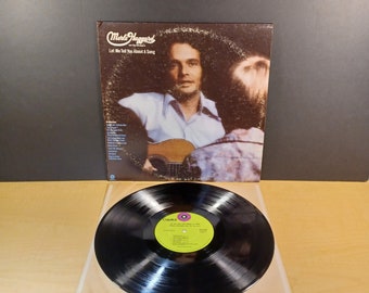 Pode incluir: Uma capa de álbum de vinil com Merle Haggard e os Strangers. A capa é marrom com uma foto de Merle Haggard segurando um violão. O título do álbum é "Let Me Tell You About A Song".