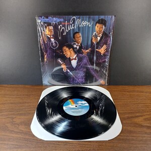 May include: A vinyl record and album cover featuring the group New Edition. The album cover has the words "Blue Moon" in script. The record is black with a colorful label. The album cover shows the group in purple sequined jackets.