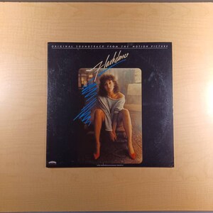 May include: A black vinyl record album cover with the title "Flashdance" in white lettering. The cover features a woman in a white sweater and red shoes sitting on a chair. The album is the original soundtrack from the motion picture.