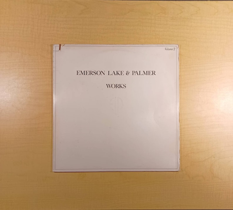Puede incluir: Un &aacute;lbum de vinilo blanco con el texto "Emerson Lake & Palmer" y "Works" impreso en marr&oacute;n en la portada. El texto "Volumen 2" est&aacute; impreso en marr&oacute;n en la esquina superior derecha.