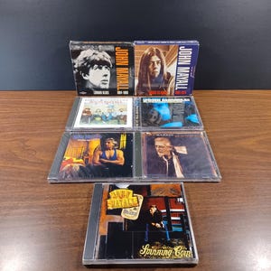 May include: A collection of John Mayall CDs, featuring album covers with portraits and text. The CDs are in clear plastic cases, with titles like "London Blues" and "Spinning Coin" visible. The image is taken on a wooden surface.