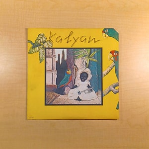 May include: A yellow record album cover with the word "Kalyan" written in black cursive. The cover features a colorful illustration of a child in a white dress holding a trumpet, surrounded by tropical plants and parrots.