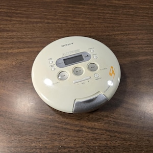 May include: A round, cream-colored Sony CD Walkman portable music player featuring an MP3 logo, a digital display screen, and various control buttons including play, pause, volume, and track skip. The device has a silver latch at the bottom.