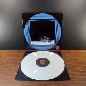 May include: A white vinyl record with a black and white image of a building on the cover. The record is titled "The Walt" and is on a black turntable mat.