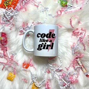 May include: A white coffee mug with the text "code like a girl" in black and pink. The mug is surrounded by candy and pink and white paper shreds on a white fluffy surface.