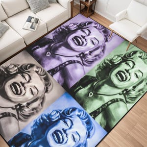 Puede incluir: Alfombra rectangular con un retrato de mujer de estilo pop art en cuatro colores diferentes: gris, morado, verde y azul. La alfombra está colocada sobre un suelo de madera.