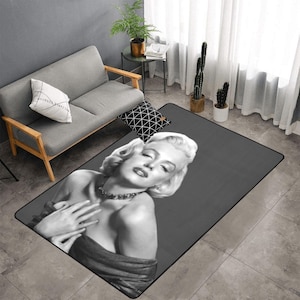 Puede incluir: Una alfombra rectangular con una imagen en blanco y negro de Marilyn Monroe. La alfombra tiene un fondo gris oscuro y un borde negro. La imagen muestra a Monroe con los ojos cerrados y las manos en el pecho.