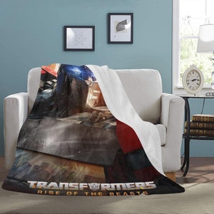 May include: A white fleece blanket with a Transformers: Rise of the Beasts movie poster printed on it. The poster features a robot in a battle pose with a blue and red color scheme.
