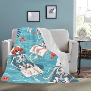 May include: A turquoise blanket featuring a whimsical skeleton pool party design. The blanket depicts skeletons relaxing in a pool with orange hats and flowers. The blanket is draped over a white couch.
