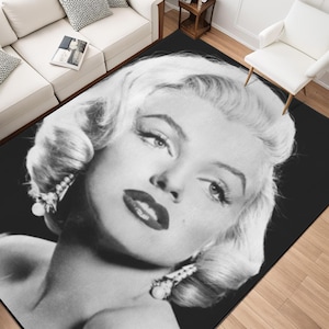 May include: Black and white area rug featuring a close-up portrait of Marilyn Monroe. The rug showcases her iconic face with bold red lipstick, styled blonde hair, and pearl earrings. The background is solid black.