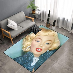 Puede incluir: Alfombra rectangular con un primer plano del rostro de Marilyn Monroe. La alfombra tiene un fondo azul claro con un borde negro. El pelo rubio, el lápiz labial rojo y los ojos azules de Monroe se muestran de forma destacada.