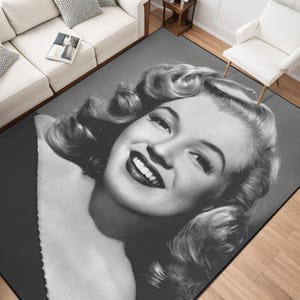 Puede incluir: Una alfombra en blanco y negro con un retrato sonriente de una mujer con cabello rizado. La alfombra tiene un fondo gris y está colocada en un salón con muebles blancos y suelo de madera.