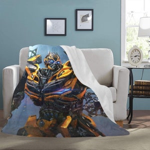 Puede incluir: Un robot amarillo y negro con ojos azules, de pie sobre un fondo azul y gris. El robot es un personaje popular de la franquicia de películas Transformers.