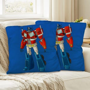 Puede incluir: Dos cojines cuadrados azules con una ilustración de dibujos animados de Optimus Prime. El robot está representado en rojo, blanco y azul, con detalles amarillos. Los cojines están sobre un sofá blanco.